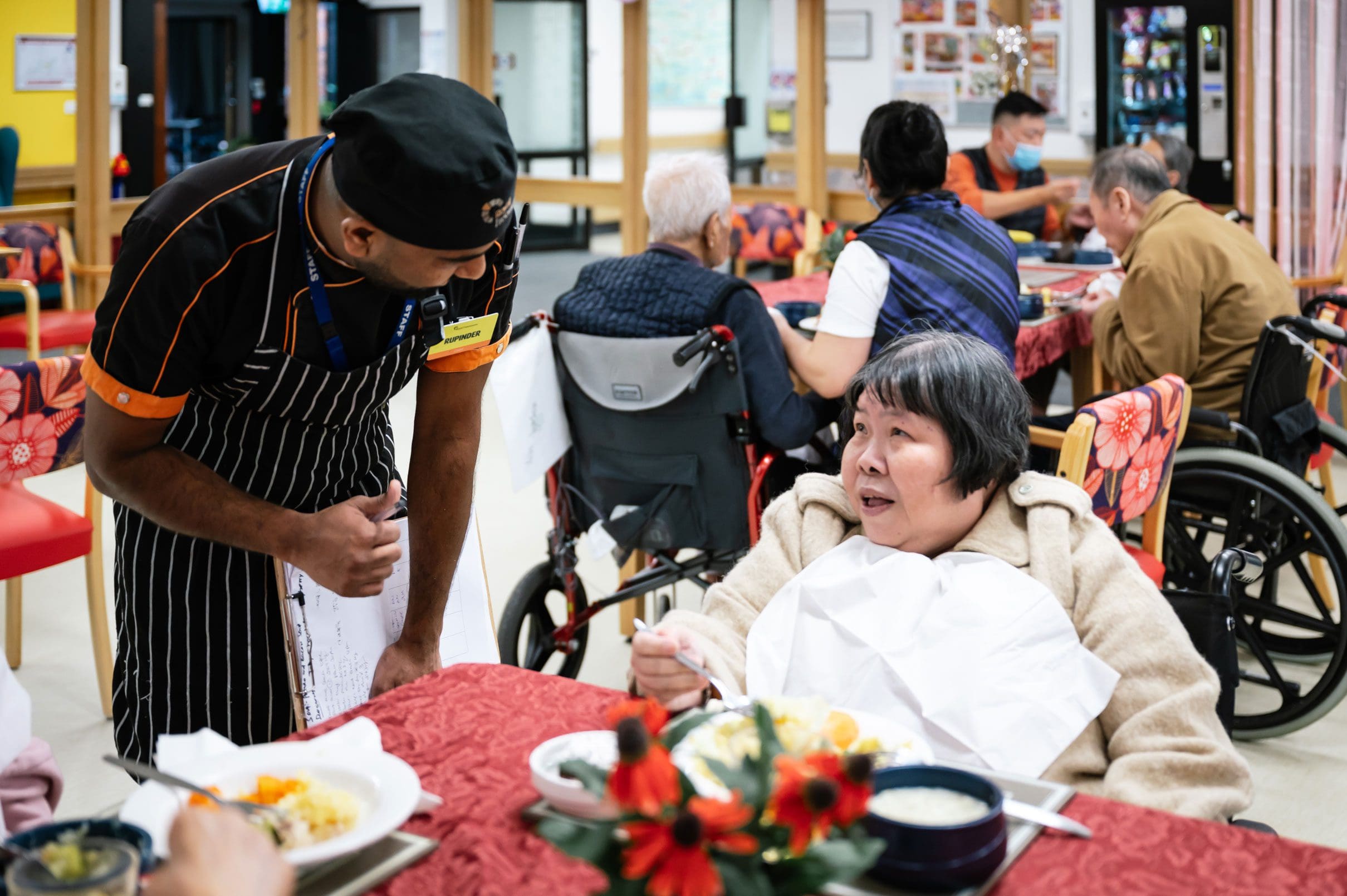 A woman in a wheelchair sitting at a table with a waiter in a retirement home.