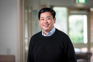 A smiling man wearing a black sweater and blue collared shirt indoors.