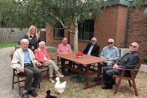 A group of elderly individuals sitting at an outdoor table with a woman standing behind them, while a duck is present in the foreground.