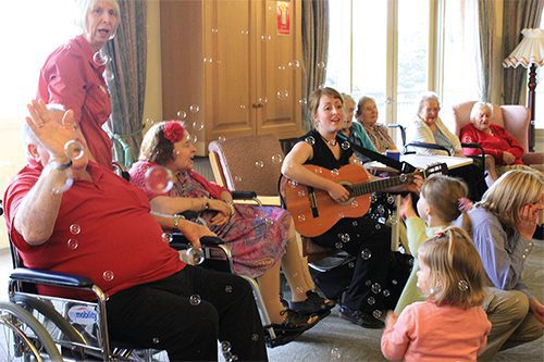A group of seniors and children enjoying a musical performance in a living room setting, with bubbles floating in the air.