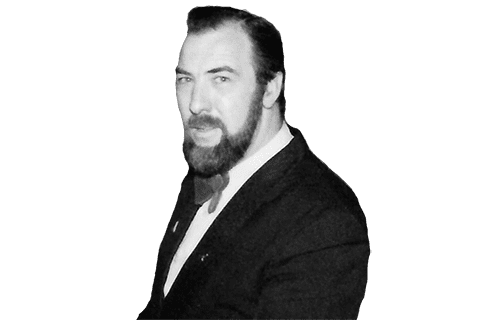 A bearded man in a dark suit standing against a green background.