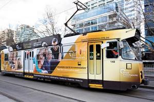 A tram covered in advertising wraps, traveling through an urban area.