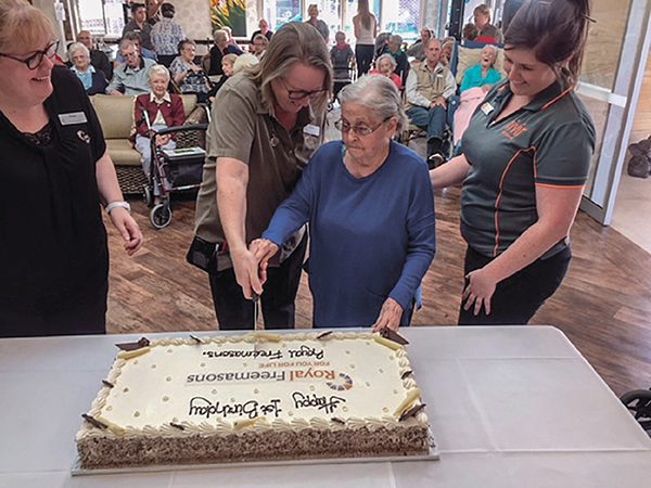 Two women cutting a large sheet cake at an event with onlookers in the background.
