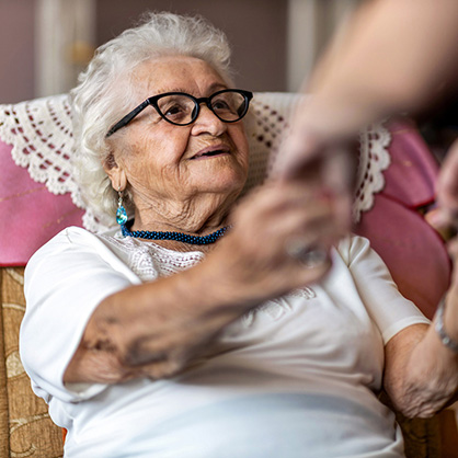 Female home carer supporting old woman to stand up from the armchair at care home
