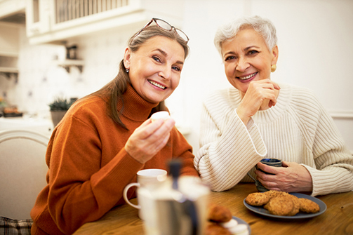 People, friendship, age and retirement concept. Indoor shot of two elegant happy women pensioners sitting at cafe table, drinking coffee and eating cookies during meeting, having nice conversation