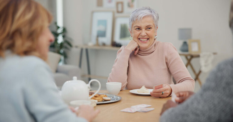 Tea, happy and retirement with friends and playing cards in living room for relax, diversity and poker. Games, smile and community with group of old people in nursing home for party and celebration
