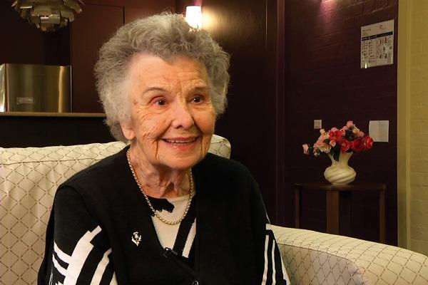 An elderly woman with a warm smile sitting on a sofa in a room with a vase of flowers in the background.