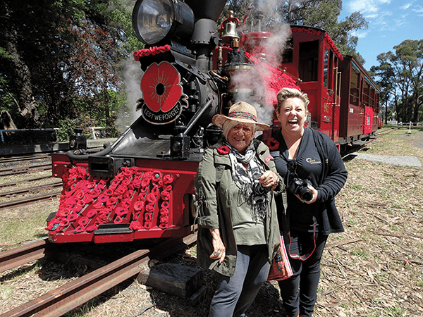 Two joyful tourists posing in front of a vintage red steam locomotive with remembrance poppies.