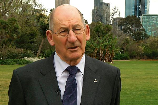 An older man in a suit with a tie and a pin on the lapel speaking outdoors with trees and city buildings in the background.
