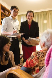 Three people delivering documents to an elderly woman sitting in a chair.
