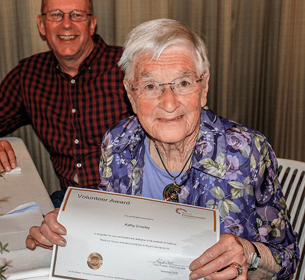 An elderly person holding a volunteer award certificate, smiling at the camera, with a standing man in the background.