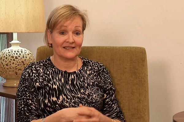 A woman with short blond hair, wearing a patterned top, seated on a green couch and conversing.