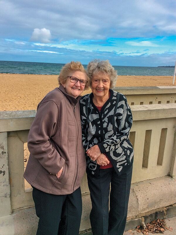 Two elderly women smiling and standing together by a seaside railing with a beach and overcast sky in the background.