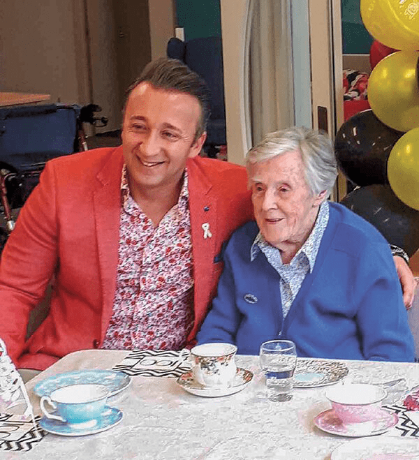 A man in a red blazer sitting beside an elderly woman in a blue cardigan at a table with tea cups, both smiling.