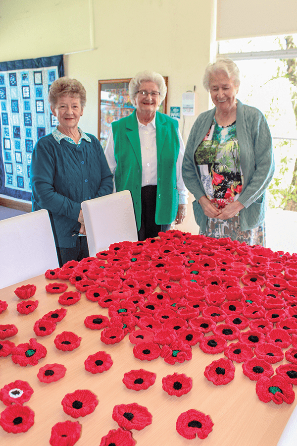 Three women standing beside a table covered with red knitted poppies.